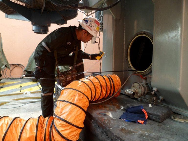 Technician checking gas levels at a tank manway with an explosion-proof ventilation duct during FPSO tank entry